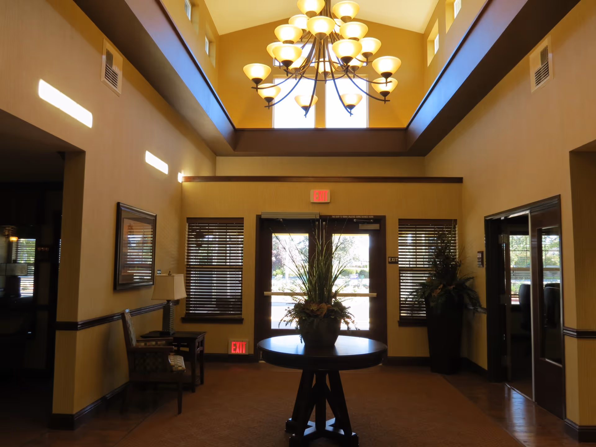Interior view of a well-lit lobby area with a high ceiling and a large chandelier. There is a round table with a floral arrangement in the center, two windows with blinds on either side of double glass exit doors, and seating with a lamp on the left side. The walls are painted yellow with dark wood trim.