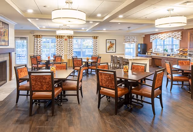 Bright, well-lit dining room with multiple wooden tables and upholstered chairs, pendant lights, and an open kitchen/service area.