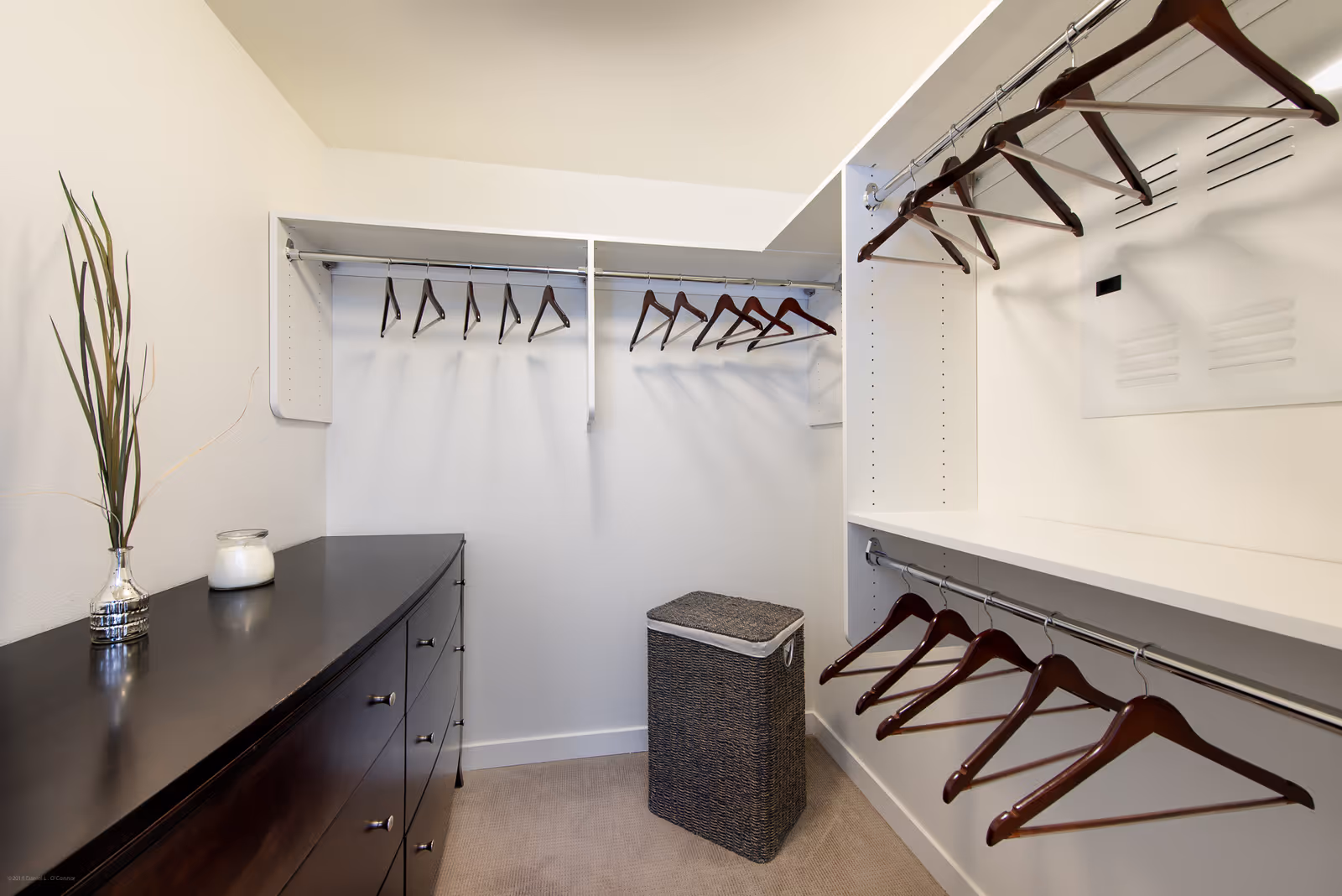 A walk-in closet with white walls and beige carpet. The closet features multiple hanging rods with wooden hangers, a dark wooden dresser with drawers on the left side, a woven laundry basket in the center, and a decorative vase with tall dried plants and a candle on top of the dresser.