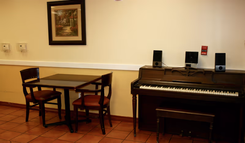 A small seating area with a wooden table and two chairs next to an upright piano with a bench. There is a framed painting on the wall above the table, and the floor is tiled with reddish-brown tiles.
