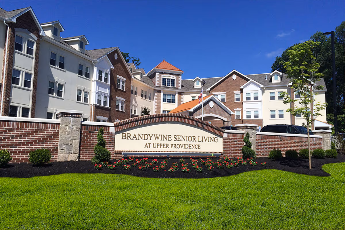 Exterior view of Brandywine Senior Living at Upper Providence building with a brick and stone sign in front, surrounded by green grass, small bushes, and flowers under a clear blue sky.