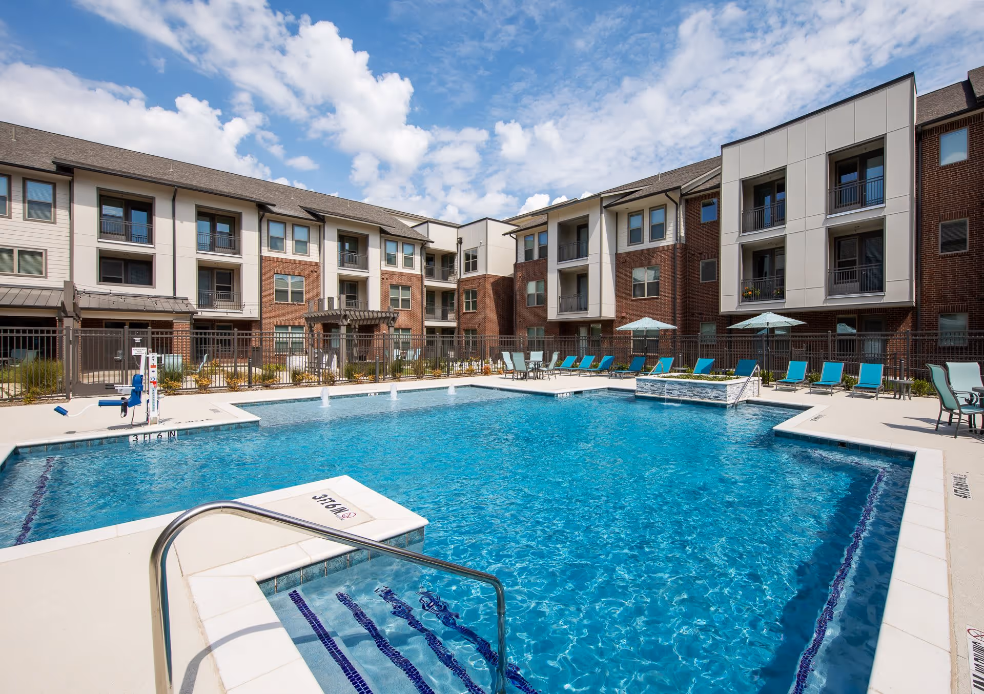 Outdoor swimming pool with clear blue water surrounded by lounge chairs and umbrellas, set in the courtyard of a multi-story residential building under a partly cloudy sky.