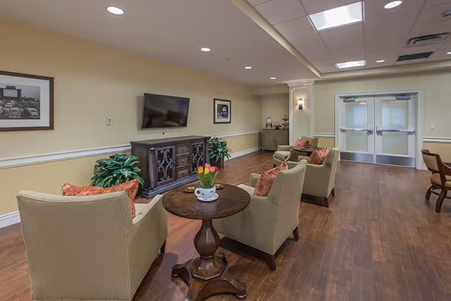 A cozy living room area in a senior living facility with four beige armchairs arranged around a round wooden table with a vase of tulips. A flat-screen TV is mounted on the wall above a wooden cabinet. The room has wood flooring, soft yellow walls with white trim, framed black and white photos, and double doors at the far end.
