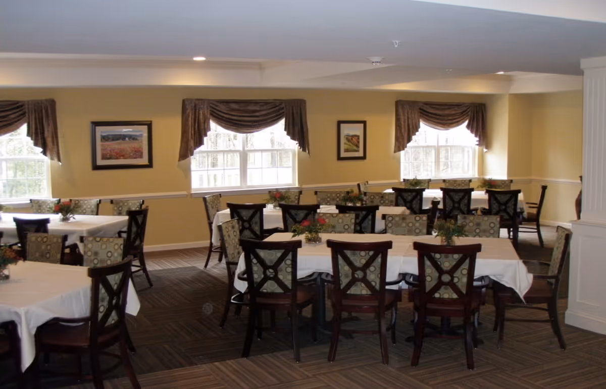 Spacious dining room with multiple tables and chairs set with white tablecloths and small floral centerpieces beneath windows with draped valances.