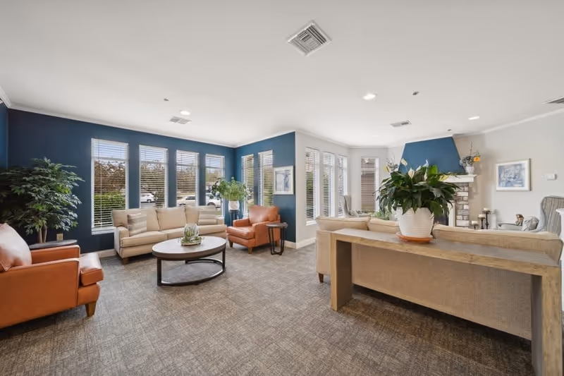 A bright and spacious living room area with large windows allowing natural light to fill the space. The room features beige sofas, two orange armchairs, a round coffee table with a decorative plant, and a wooden console table with a large potted plant. The walls are painted blue and white, and there is a fireplace with a blue accent wall in the corner. The carpet is patterned in neutral tones.