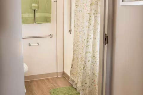 View into a bathroom featuring a shower with a floral-patterned curtain, a green bath mat on the floor, green towels hanging on a rack, and a grab bar on the wall.