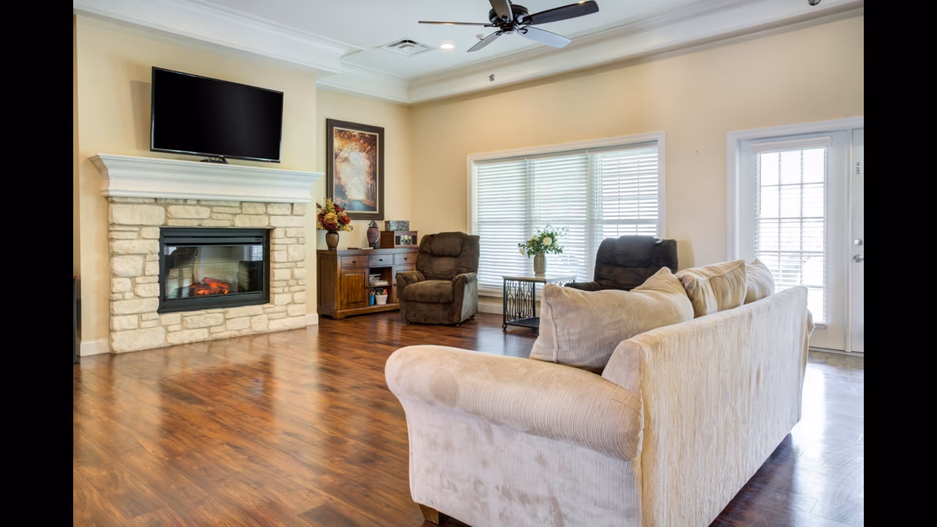 Bright living room with a stone fireplace and wall-mounted TV, a sofa and recliners on hardwood floors beneath a ceiling fan.