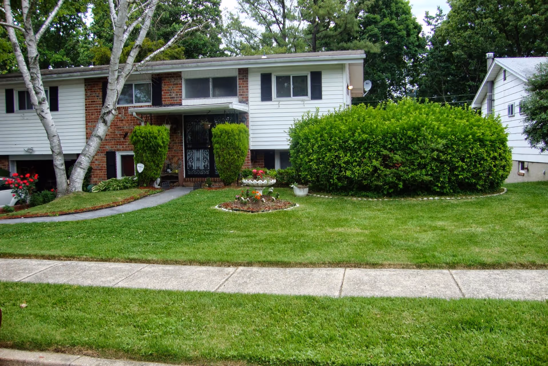 Front exterior view of a two-story house with white siding and brick accents, surrounded by green grass, bushes, and trees. A concrete sidewalk runs in front of the house, and a small flower bed is visible near the entrance.