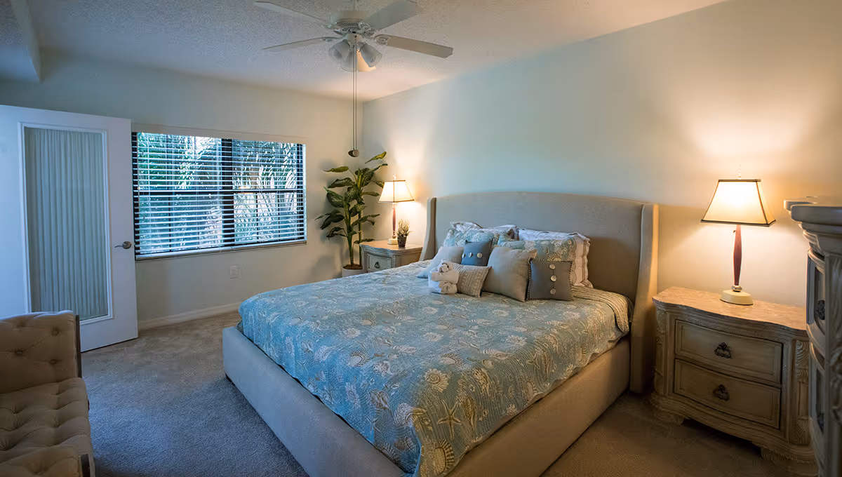 A cozy bedroom with a large bed covered in a blue patterned bedspread and multiple pillows. There are two wooden nightstands on either side of the bed, each with a lamp. A ceiling fan is mounted above, and a window with blinds lets in natural light. A potted plant is placed near the window, and a cushioned chair is partially visible in the corner.