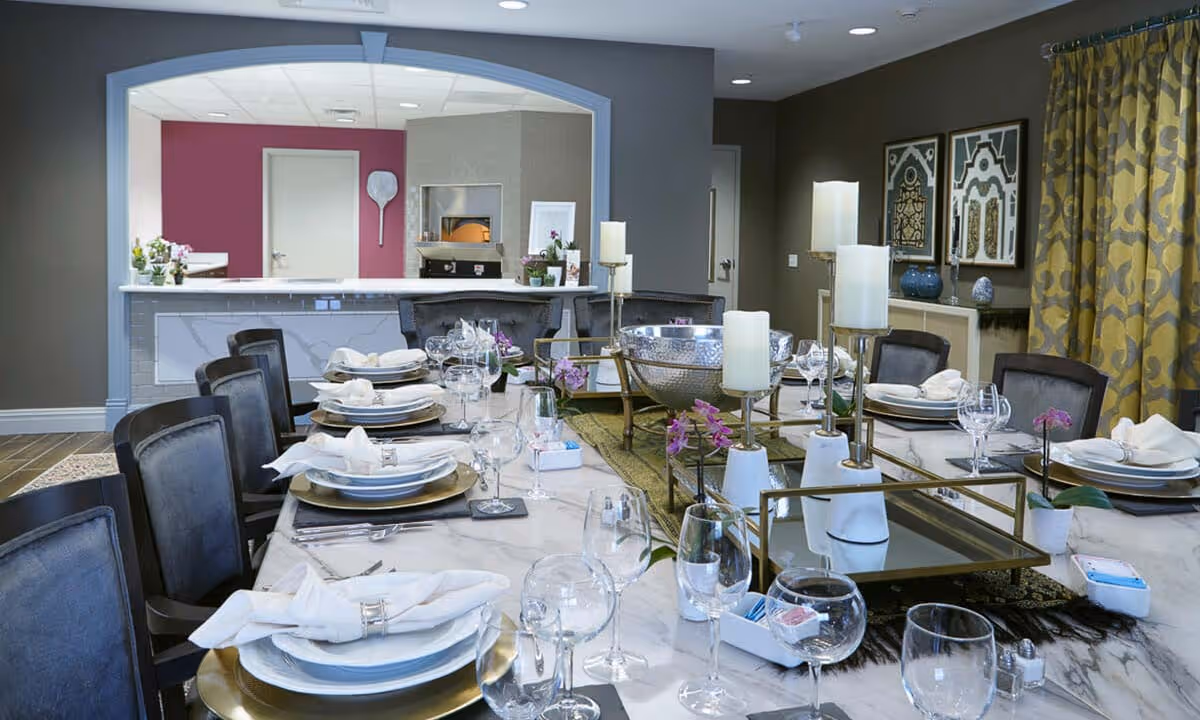 A formal dining room with a long marble table set for a meal, featuring plates, napkins, wine glasses, and silverware. The table is decorated with candles, small purple flowers, and a large silver bowl centerpiece. The room has dark walls, patterned yellow curtains, framed artwork, and a serving counter with a pizza oven in the background.