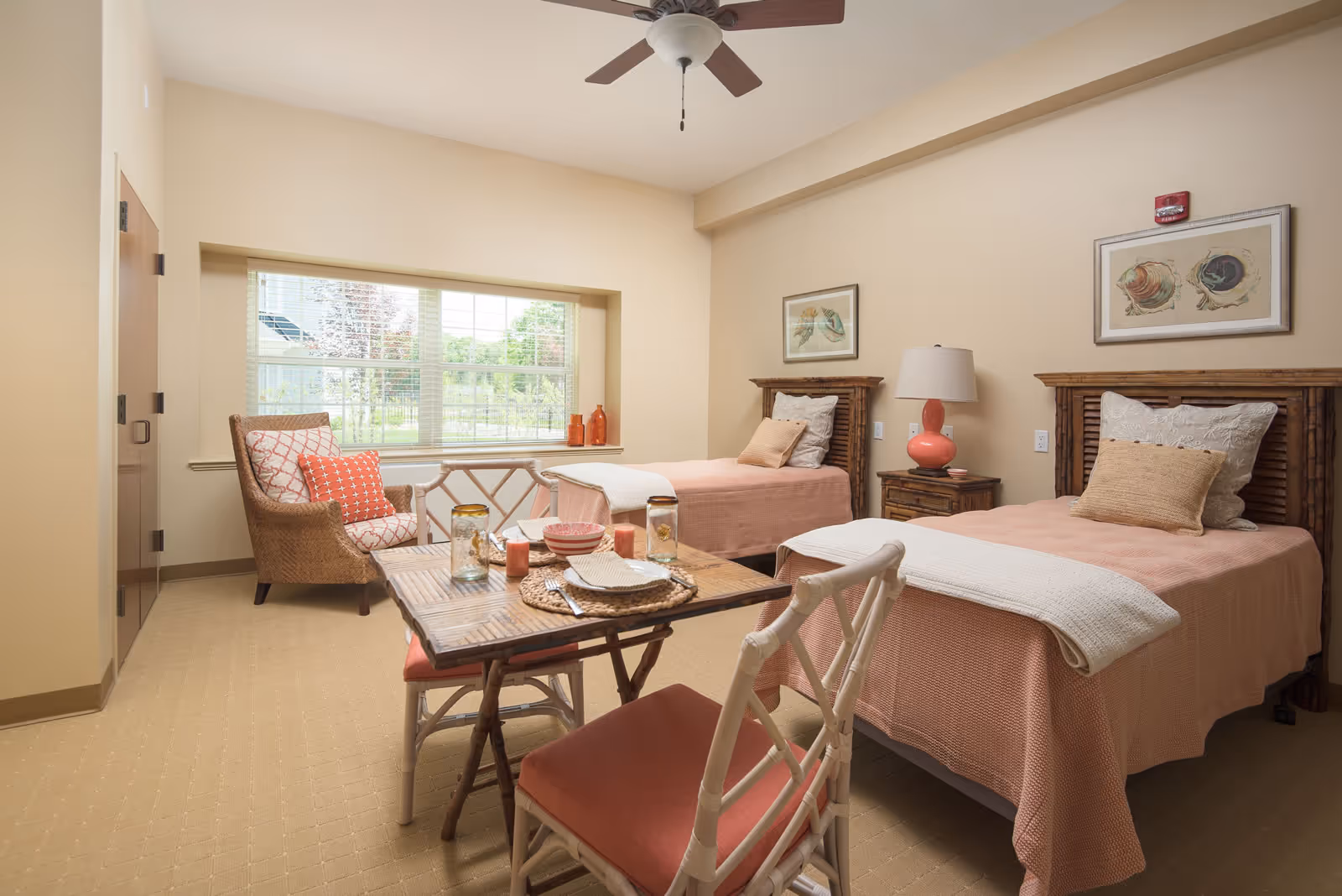 A bright senior living bedroom with two twin beds covered in peach-colored bedspreads and white pillows. Between the beds is a wooden nightstand with a coral-colored lamp. A wooden table with two chairs is in the foreground, set with dishes and candles. A wicker armchair with patterned cushions sits near a large window that lets in natural light. The walls are painted beige and decorated with framed artwork.