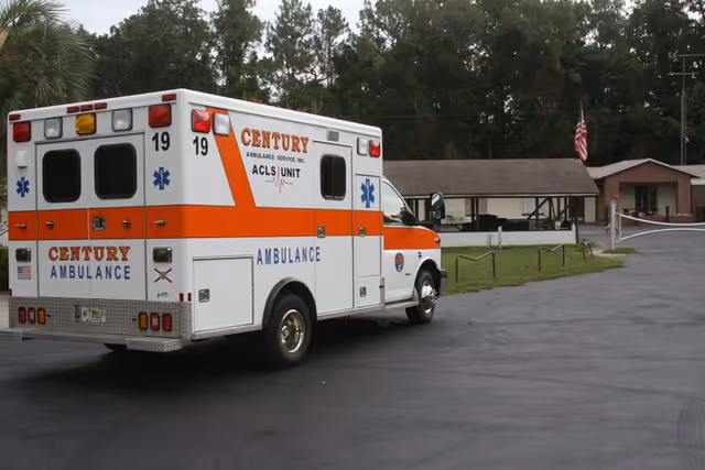 An ambulance parked in front of a single-story building with an American flag and trees in the background.