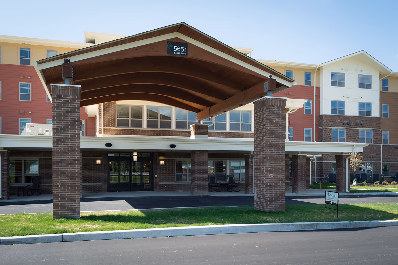 Front entrance of a multi-story senior living facility with a large covered driveway supported by brick pillars. The building has a mix of brick and light-colored siding with multiple windows. A sign near the entrance reads 'Oasis At 30th'.