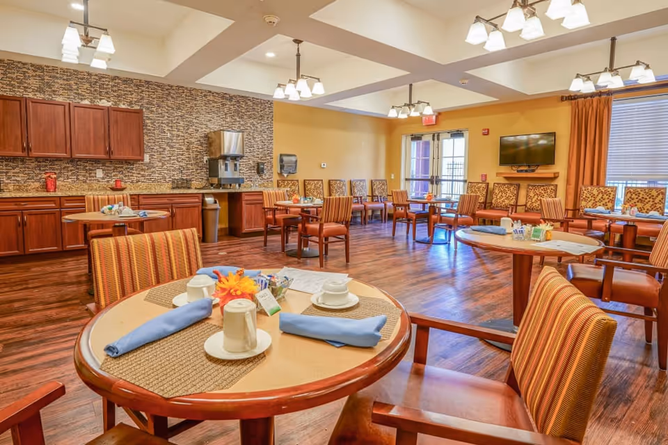 A bright dining room with round wooden tables set with cups, saucers, and blue napkins. The room features wooden chairs with striped upholstery, a wall-mounted TV, large windows with orange curtains, and a counter area with cabinets and a beverage dispenser. The floor is wooden, and the walls are painted yellow with a textured backsplash behind the counter.
