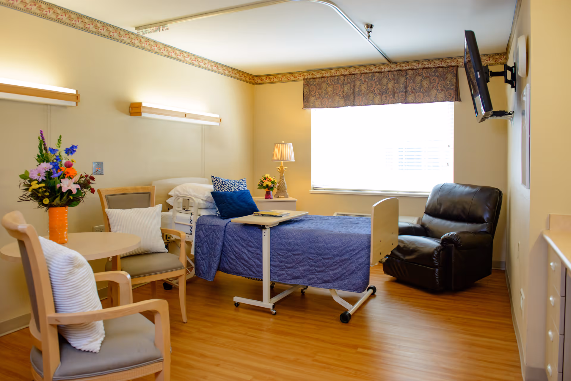 A well-lit nursing facility room with a hospital bed covered in a blue quilt, a small overbed table, a black recliner chair, a round table with two chairs, and a vase of colorful flowers. The room has wooden flooring, a window with a patterned valance, and a wall-mounted TV.