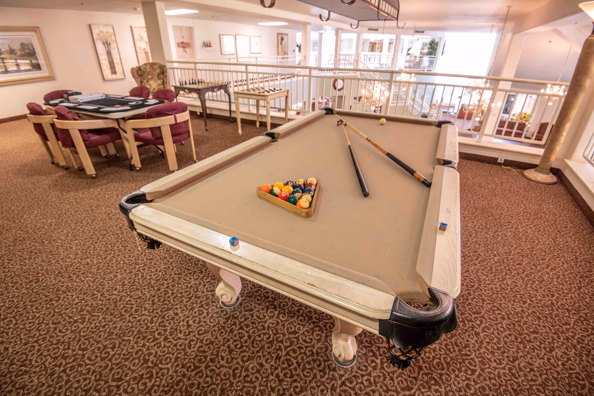 A senior living facility game room featuring a beige pool table with pool balls racked and two cues resting on the table. In the background, there is a card table with four chairs and a chess set on a small table. The room has patterned carpet, framed artwork on the walls, and large windows letting in natural light.