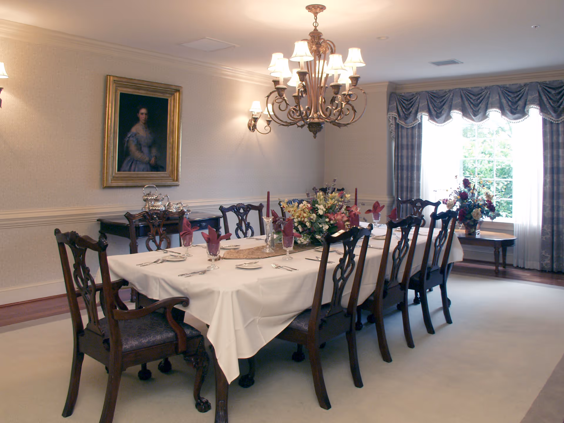 A formal dining room with a long rectangular table covered with a white tablecloth, set with plates, silverware, glasses with burgundy napkins, and floral centerpieces. The room features wooden chairs with upholstered seats, a chandelier with multiple lights, wall sconces, a portrait painting on the wall, and a large window with draped curtains allowing natural light to enter.