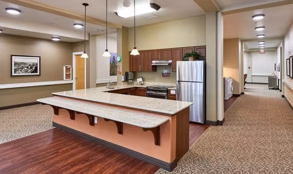 Interior view of a memory care facility kitchen area with a large marble countertop island, wooden cabinets, stainless steel refrigerator and stove, pendant lights hanging from the ceiling, and a hallway leading to other rooms.