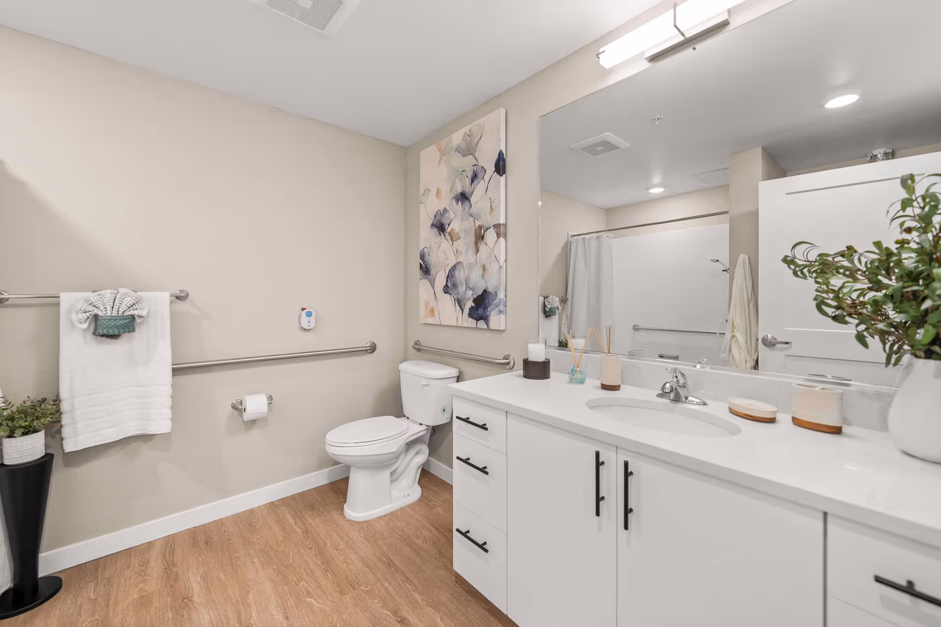 A clean and modern bathroom with a white toilet, white vanity with black handles, a large mirror, and a shower with a white curtain. The walls are beige, the floor has wood-like flooring, and there are decorative items including a plant, a towel, and a floral painting on the wall.