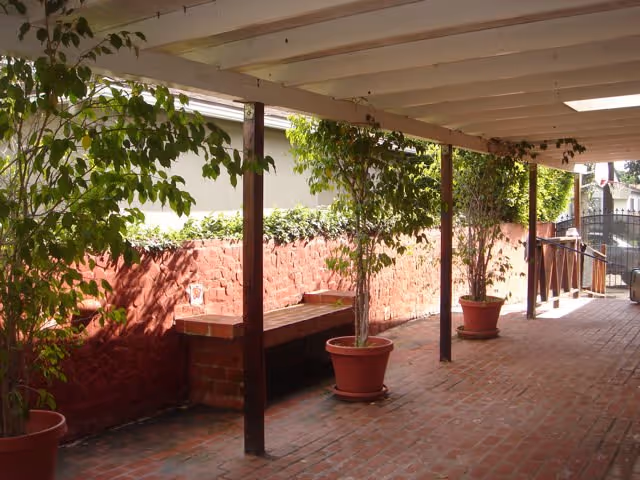 Covered outdoor walkway with brick flooring and a red brick wall on one side. Several potted plants and small trees are placed along the walkway, which is supported by wooden posts. The area is shaded by a roof with some skylights.