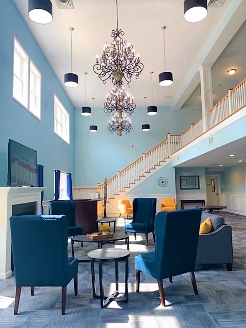 Bright two-story common lounge with teal armchairs around small tables, ornate chandeliers, a staircase and second-floor balcony.