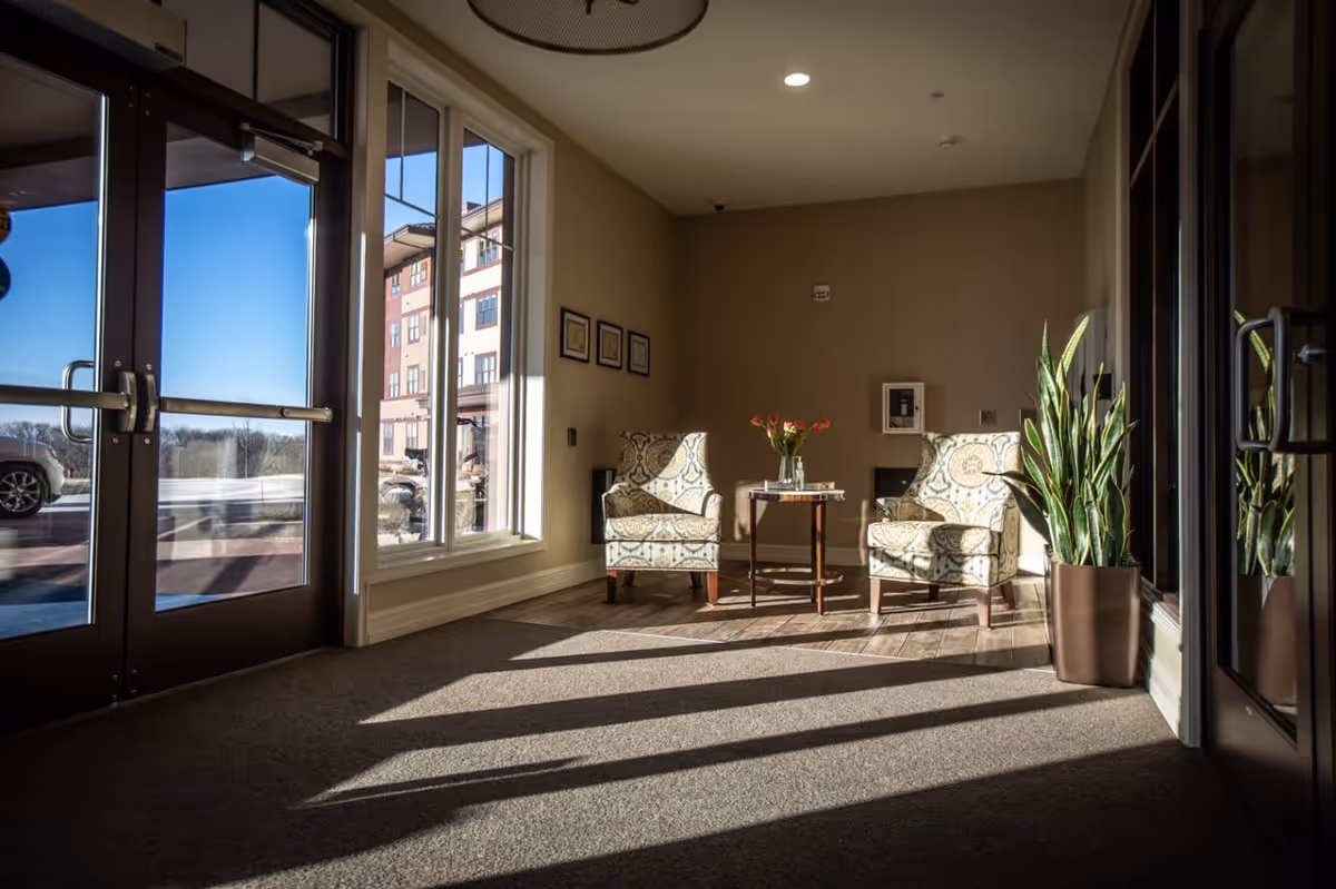 Sunlit seating area near the entrance of a facility with two patterned armchairs, a small wooden table with a vase of flowers, large windows, glass doors, and a potted plant.