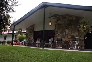 Exterior view of a building with a stone facade and a covered porch area featuring several striped cushioned chairs and potted plants, surrounded by green grass and landscaping.