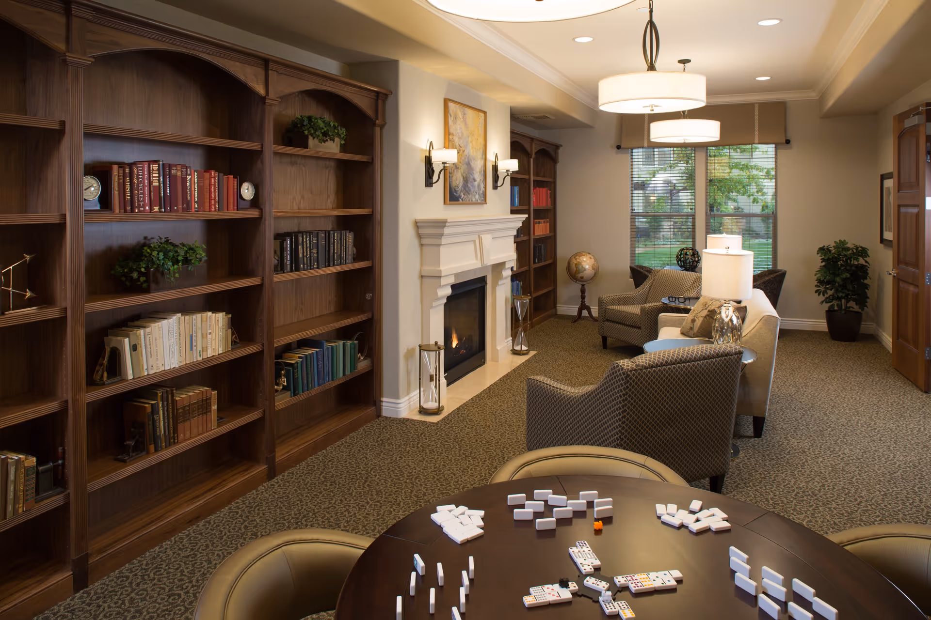 A cozy living room area in a senior living facility featuring wooden bookshelves filled with books, a fireplace with a painting above it, comfortable armchairs and a sofa, a table with dominoes set up for a game, and large windows letting in natural light.