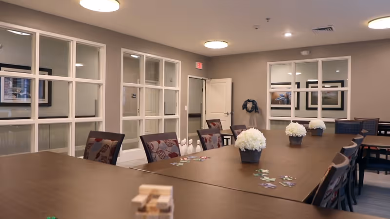 A well-lit interior room with a large wooden table surrounded by chairs. The table has small flower arrangements and some game pieces on it. The room has beige walls, large windows with white frames, and a door in the background. Ceiling lights illuminate the space.