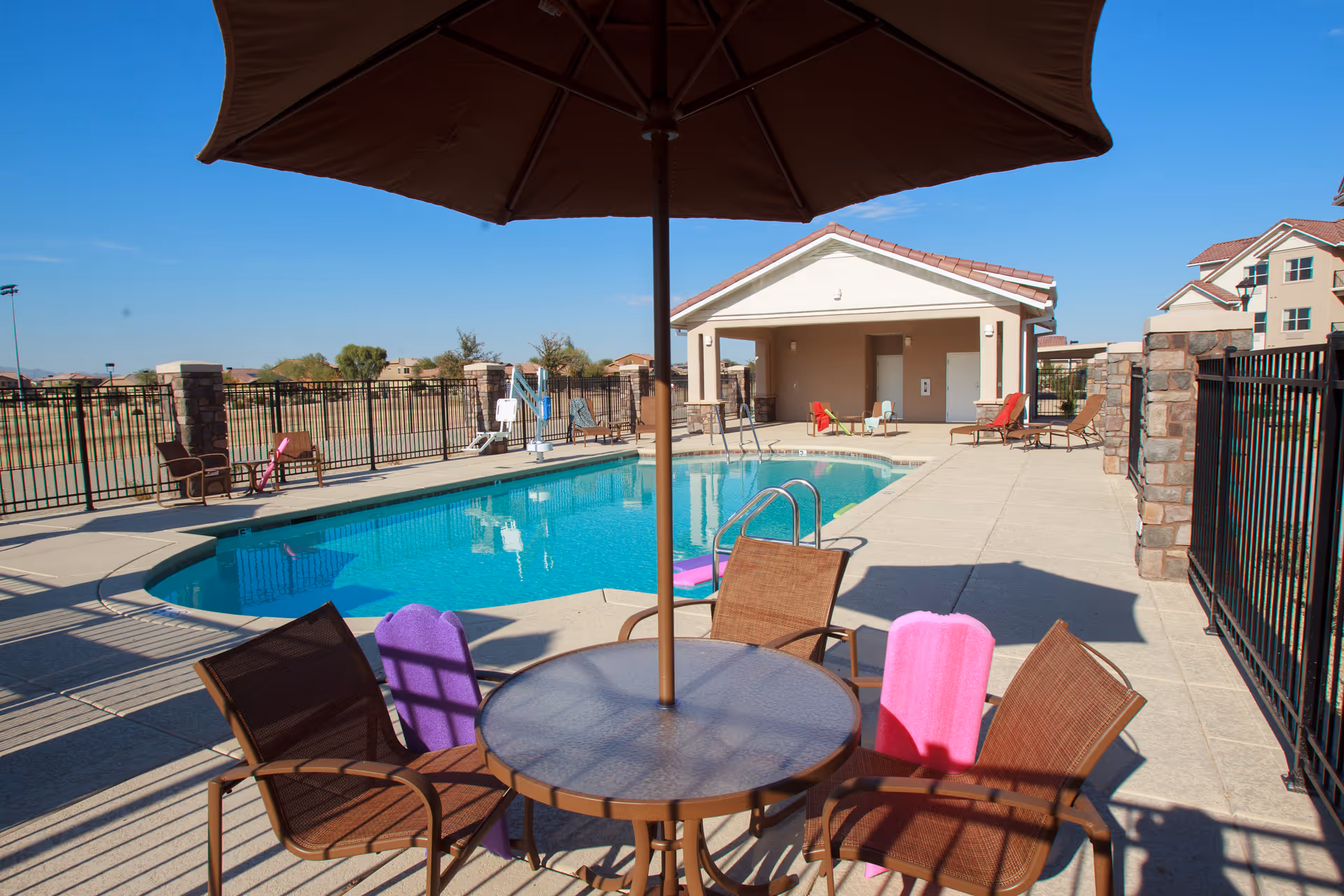 Outdoor swimming pool area with a round table and four chairs under a large umbrella. The pool is surrounded by a concrete deck with additional lounge chairs and a small building in the background. The sky is clear and blue.
