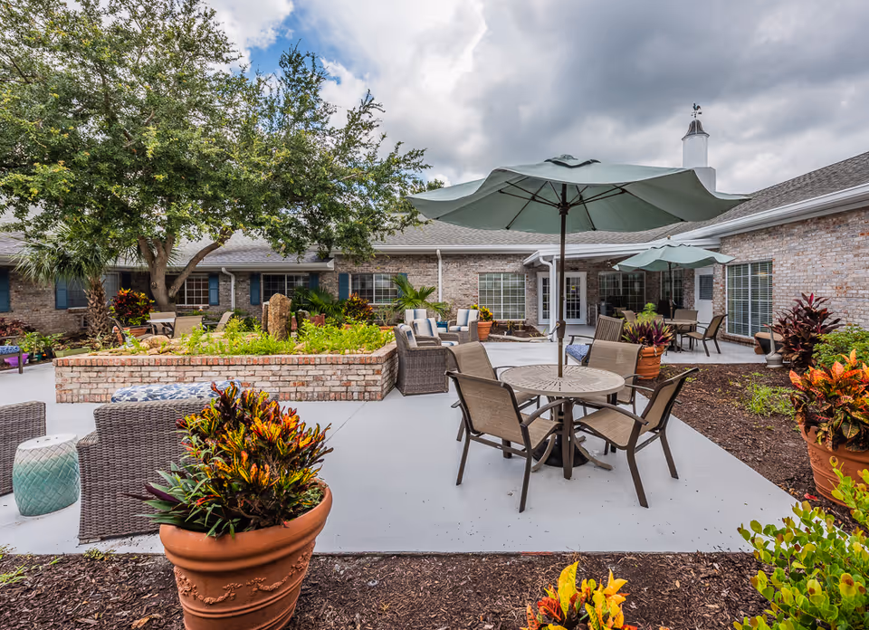 A landscaped outdoor courtyard with patio tables, umbrellas, seating and potted plants outside a brick senior living building.