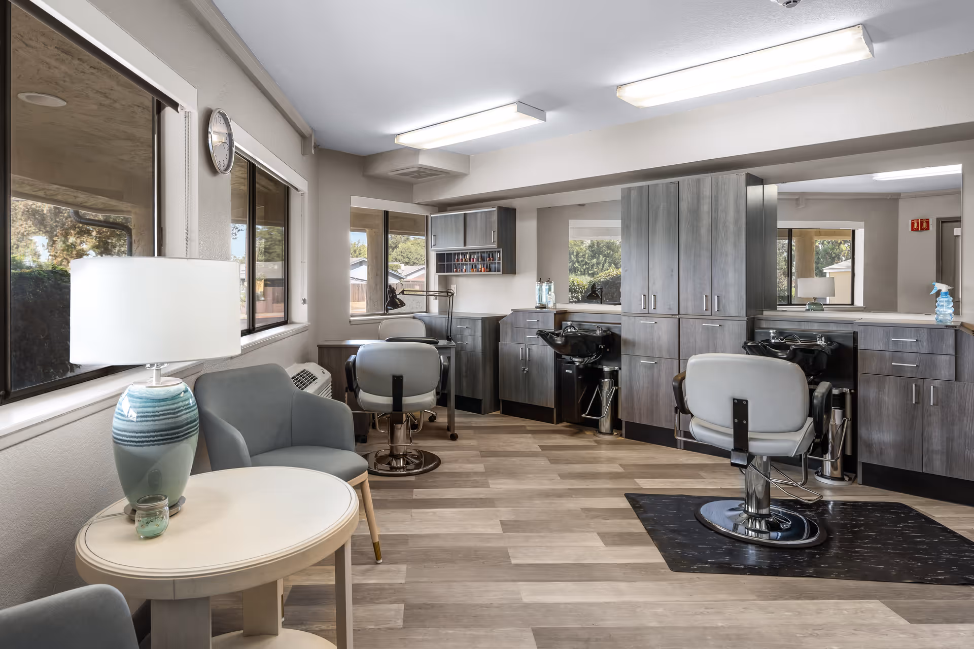 Interior view of a senior living community salon area with two salon chairs in front of wash basins, gray cabinetry, large mirrors, and a seating area with two gray armchairs and a round table with a lamp. Large windows provide natural light and a view of the outdoors.
