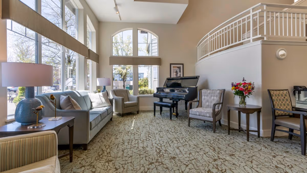 A bright and spacious living room area with large windows letting in natural light. The room features a light blue sofa, several armchairs, a black grand piano, a side table with a vase of colorful flowers, and two blue table lamps on wooden side tables. The walls are painted beige, and there is a carpet with a subtle pattern covering the floor.
