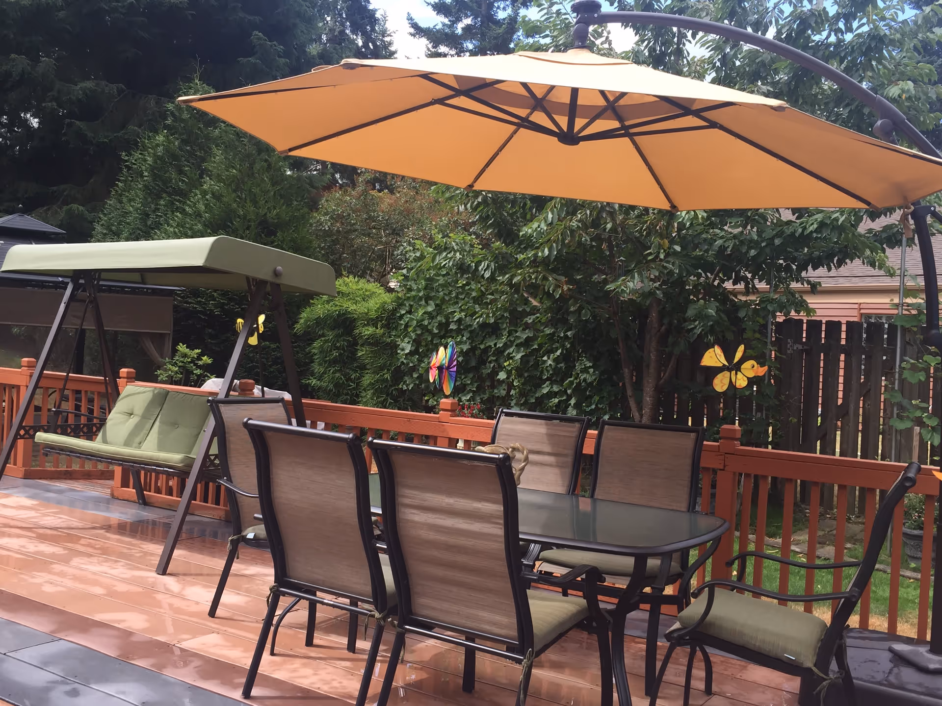 Outdoor patio area with a glass-top dining table surrounded by six chairs with beige cushions. There is a large orange umbrella providing shade over the table. To the left, there is a green cushioned swing seat. The patio is surrounded by a wooden fence and lush green trees and bushes.