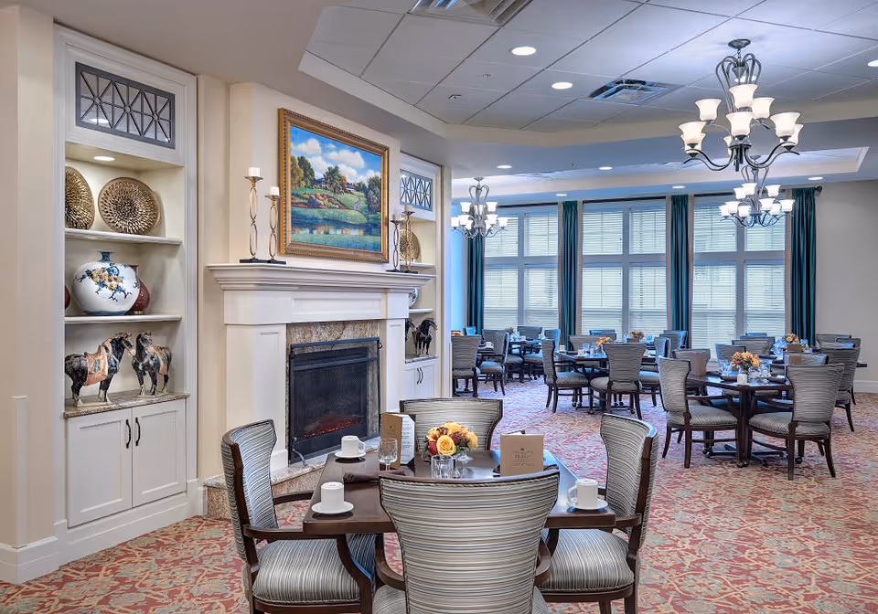 A spacious dining room in Dublin Retirement Village featuring multiple round tables with chairs arranged around them. The tables are set with cups, glasses, and small floral centerpieces. The room has large windows with blinds and dark curtains, elegant chandeliers hanging from the ceiling, and a fireplace with decorative items and a framed landscape painting above it. The carpet has a patterned design in warm tones.