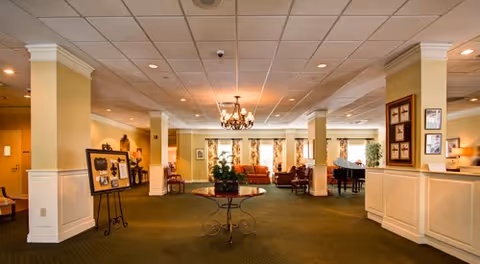 Spacious and well-lit common area in a senior living facility with green carpet, cream-colored walls, a chandelier hanging from the ceiling, and several seating areas with sofas and chairs near large windows. A round table with a plant is centered in the foreground, and framed pictures and informational boards are displayed on the walls and stands.