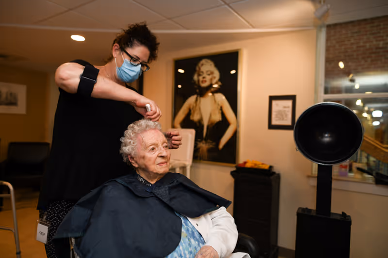 A hairstylist wearing a face mask is cutting the hair of an elderly woman seated in a salon chair. The elderly woman is covered with a black salon cape. In the background, there is a large framed picture of Marilyn Monroe on the wall and a hair dryer hood nearby.
