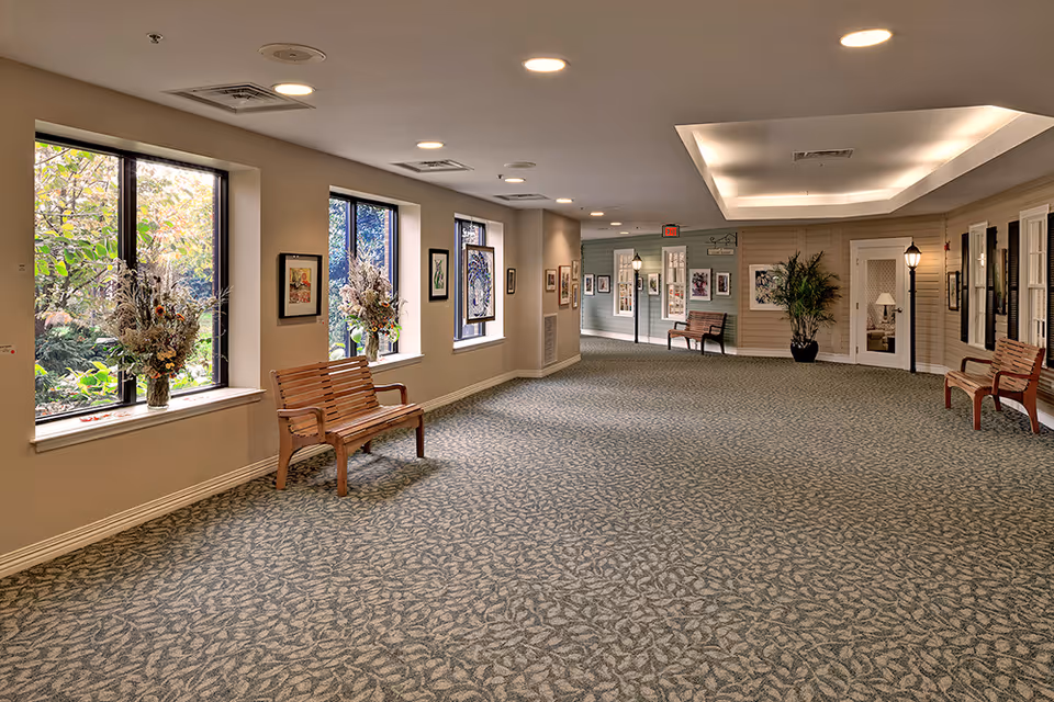 A spacious, well-lit hallway in a senior living facility with large windows on the left side adorned with flower arrangements. The hallway features patterned carpet, wooden benches along the walls, framed artwork, and decorative streetlamp-style lights. There is a door and potted plant on the right side, and an exit sign is visible in the distance.