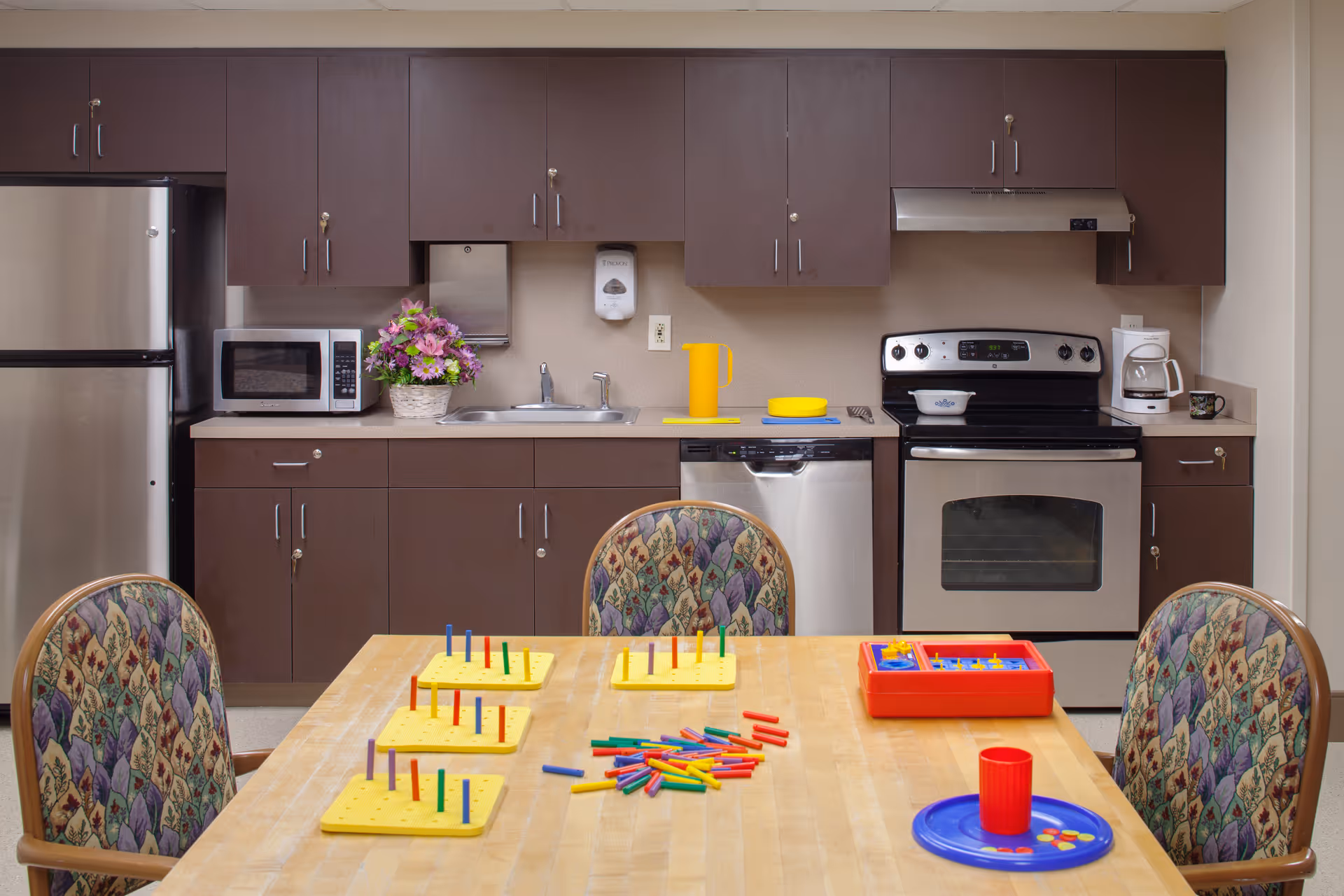Communal kitchen with dark cabinets, stainless appliances, and a table with chairs and colorful pegboard activity pieces.