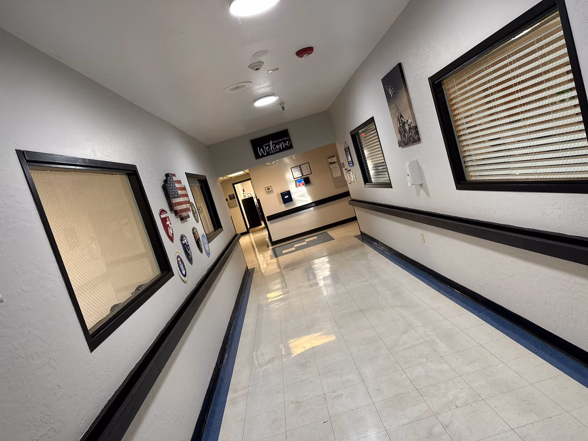 A clean, well-lit hallway in Desert Peak Care Center with white walls, black trim, and a shiny tiled floor. The hallway features windows with blinds on both sides, several plaques and decorations on the left wall, and a sign that says 'welcome' at the far end above a doorway.