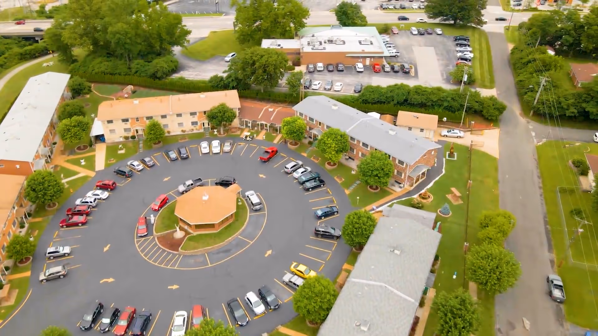 Aerial view of Florissant Valley Health and Rehabilitation facility showing multiple two-story buildings arranged around a circular driveway with parked cars. The area is surrounded by trees and greenery, with additional parking lots and roads visible in the background.