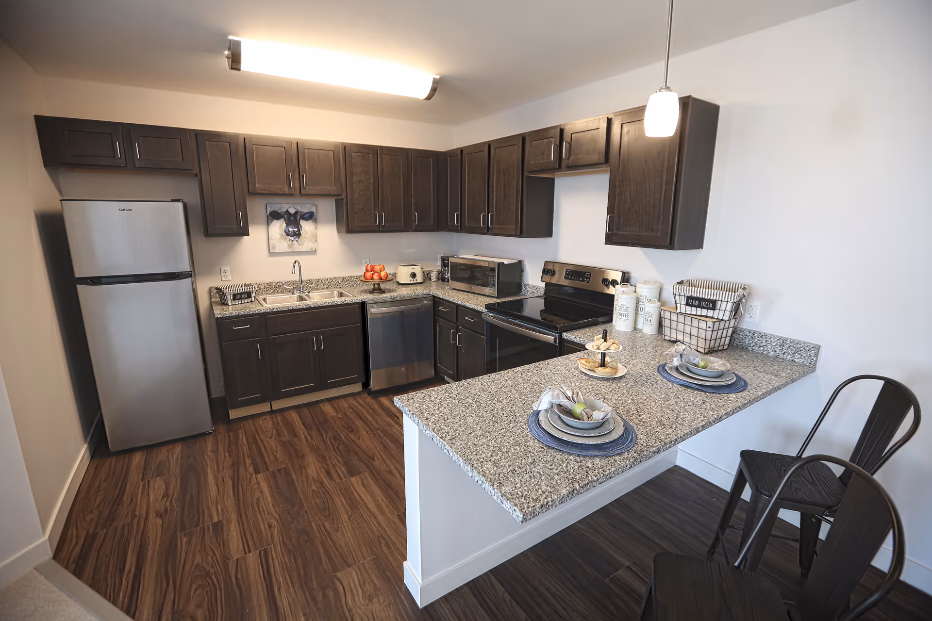 Modern kitchen with dark wood cabinets, stainless steel refrigerator, dishwasher, and stove. Granite countertops with a breakfast bar set with two place settings and two black metal chairs. Wooden floor and a wall-mounted light fixture above the sink area.