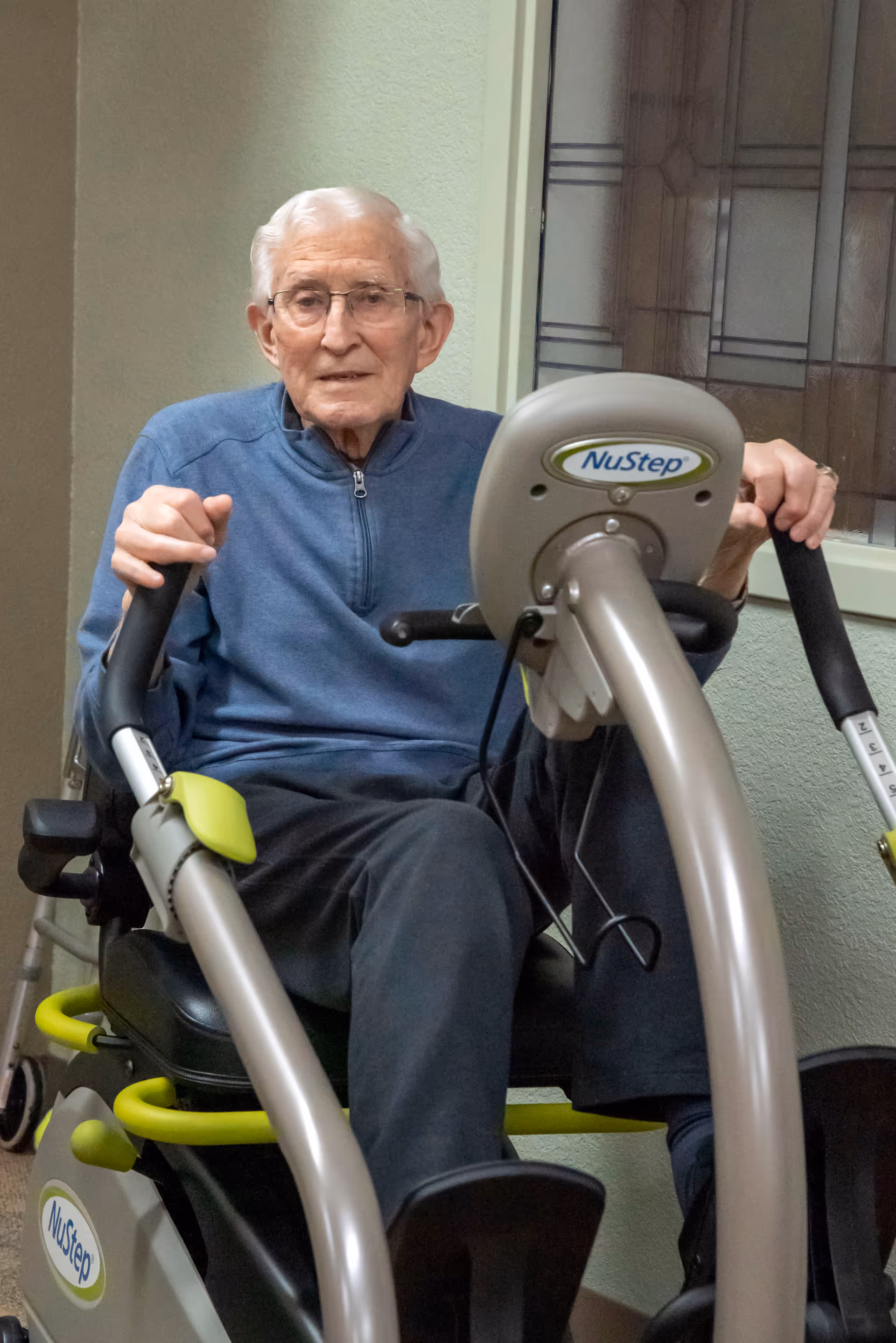 An elderly man wearing glasses and a blue zip-up sweater is seated on a NuStep exercise machine indoors, holding the handles and looking towards the camera.