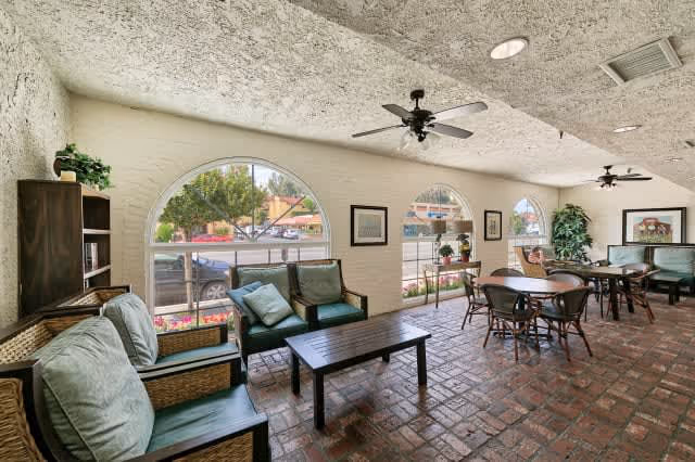 A spacious common area with brick flooring and a textured ceiling featuring ceiling fans. The room has large arched windows letting in natural light and showing a view of the street outside. There are several seating arrangements including cushioned chairs, a coffee table, and dining tables with chairs. The walls are decorated with framed pictures and there are some plants placed around the room.