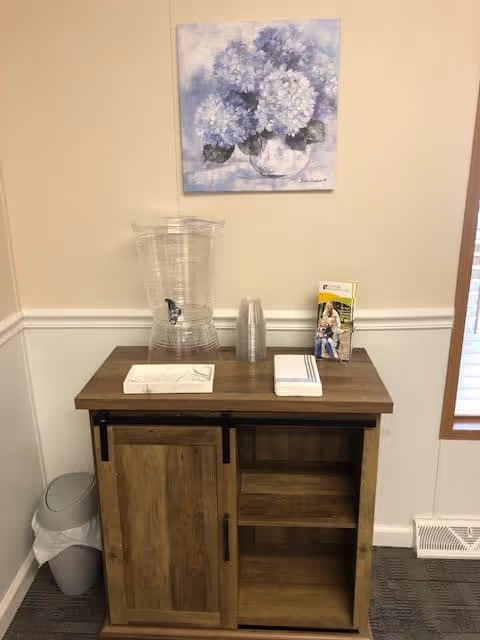 A wooden cabinet with a sliding door in a corner of a room. On top of the cabinet is a clear water dispenser with a spout, a stack of clear plastic cups, a box of tissues, a stack of napkins, and a brochure. Above the cabinet hangs a painting of blue and white hydrangea flowers in a vase. To the left of the cabinet is a trash can with a liner, and to the right is a window with blinds partially visible.