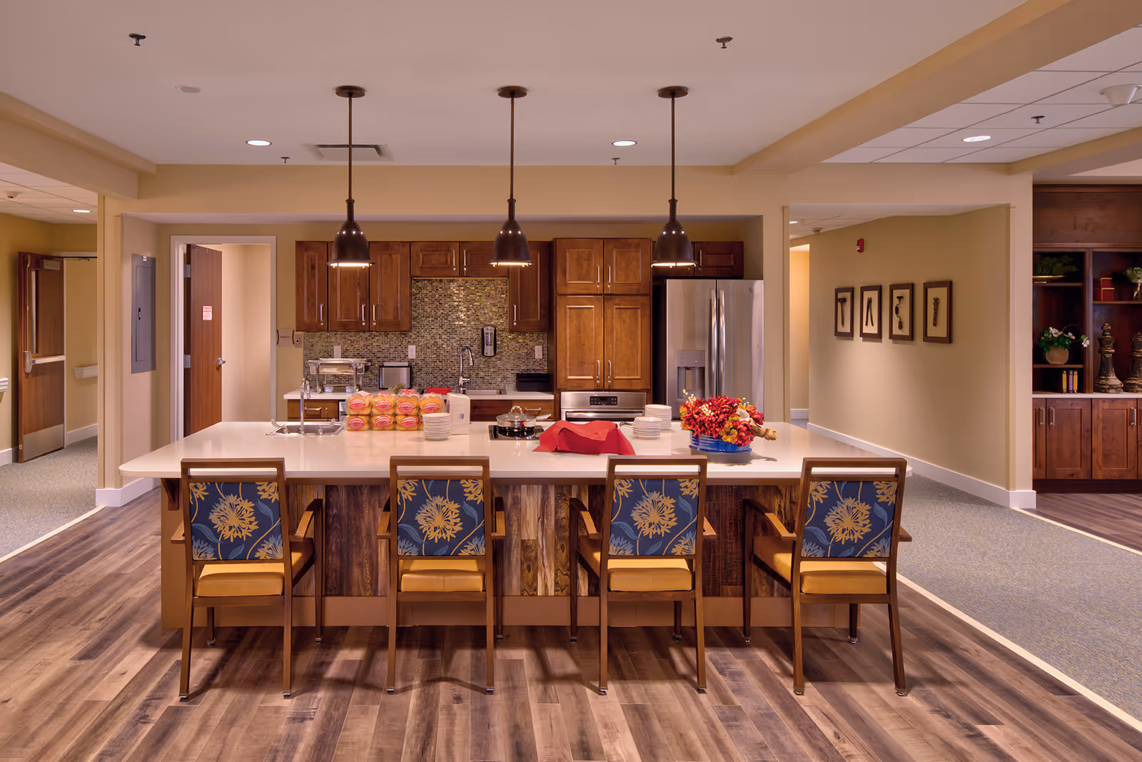 A modern kitchen area in a senior living facility with a large island countertop surrounded by four wooden chairs with floral patterned cushions. The kitchen features wooden cabinets, a stainless steel refrigerator, an oven, and a mosaic tile backsplash. Three pendant lights hang above the island, which has plates, bread rolls, a pot, and a floral centerpiece on it. The surrounding area includes wood flooring and beige walls with framed artwork.