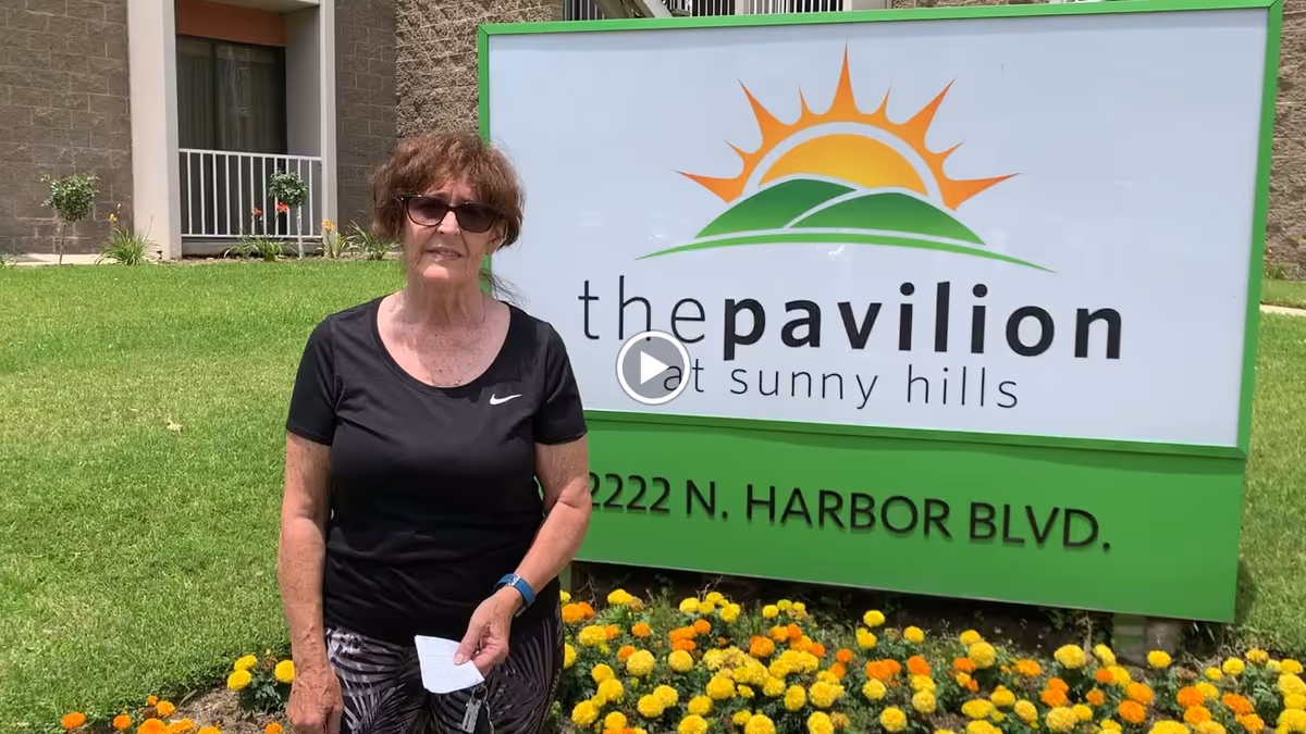 An elderly woman wearing sunglasses, a black Nike t-shirt, and patterned pants stands outside on a lawn with yellow and orange flowers in front of a large green and white sign that reads 'the pavilion at sunny hills 2222 N. Harbor Blvd.'