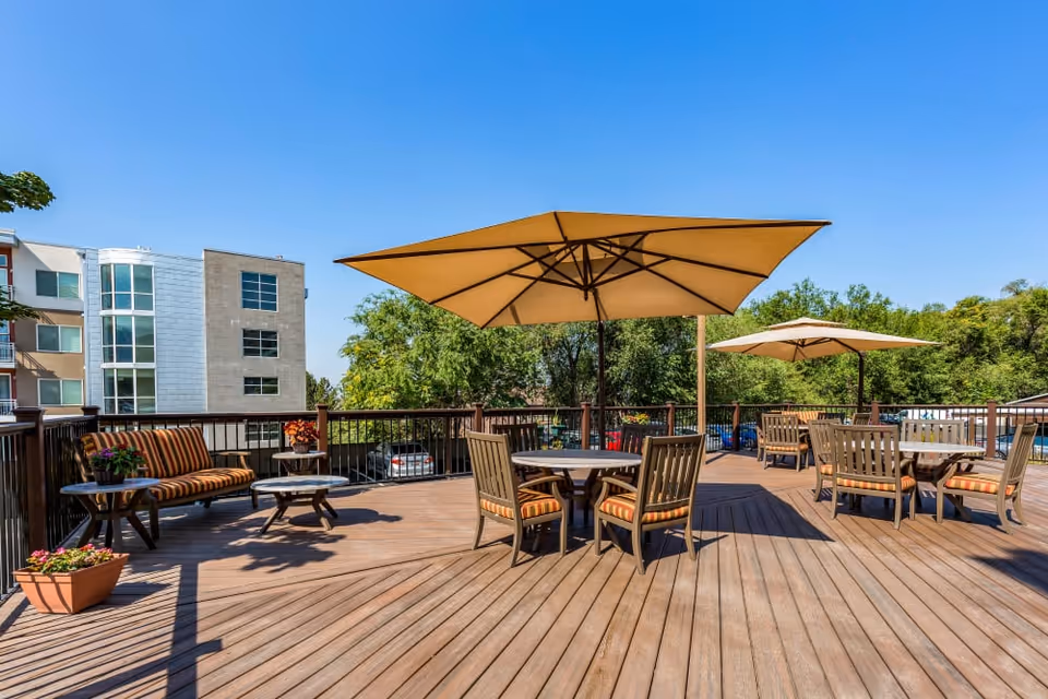 Outdoor wooden deck area with multiple round tables and chairs featuring striped cushions. Large beige umbrellas provide shade over the tables. There is a cushioned bench with a small side table and potted plants on the deck. Trees and a multi-story building are visible in the background under a clear blue sky.