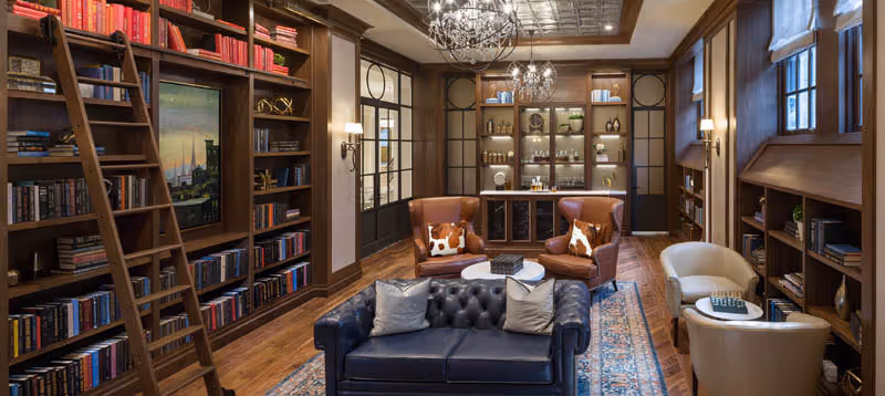 A cozy and elegant library or lounge area with wooden bookshelves filled with books, a rolling ladder, a dark blue leather tufted sofa with two pillows, two brown leather armchairs with patterned cushions, two beige armchairs, a round coffee table, and a decorative rug on a wooden floor. The room features warm lighting, a chandelier, and large windows with wooden frames and blinds.