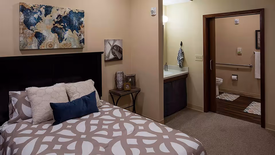 A bedroom with a bed featuring patterned bedding and multiple pillows, a small side table with decorative items, and wall art including a world map. The room opens into a bathroom area with a sink and a toilet visible through a doorway.